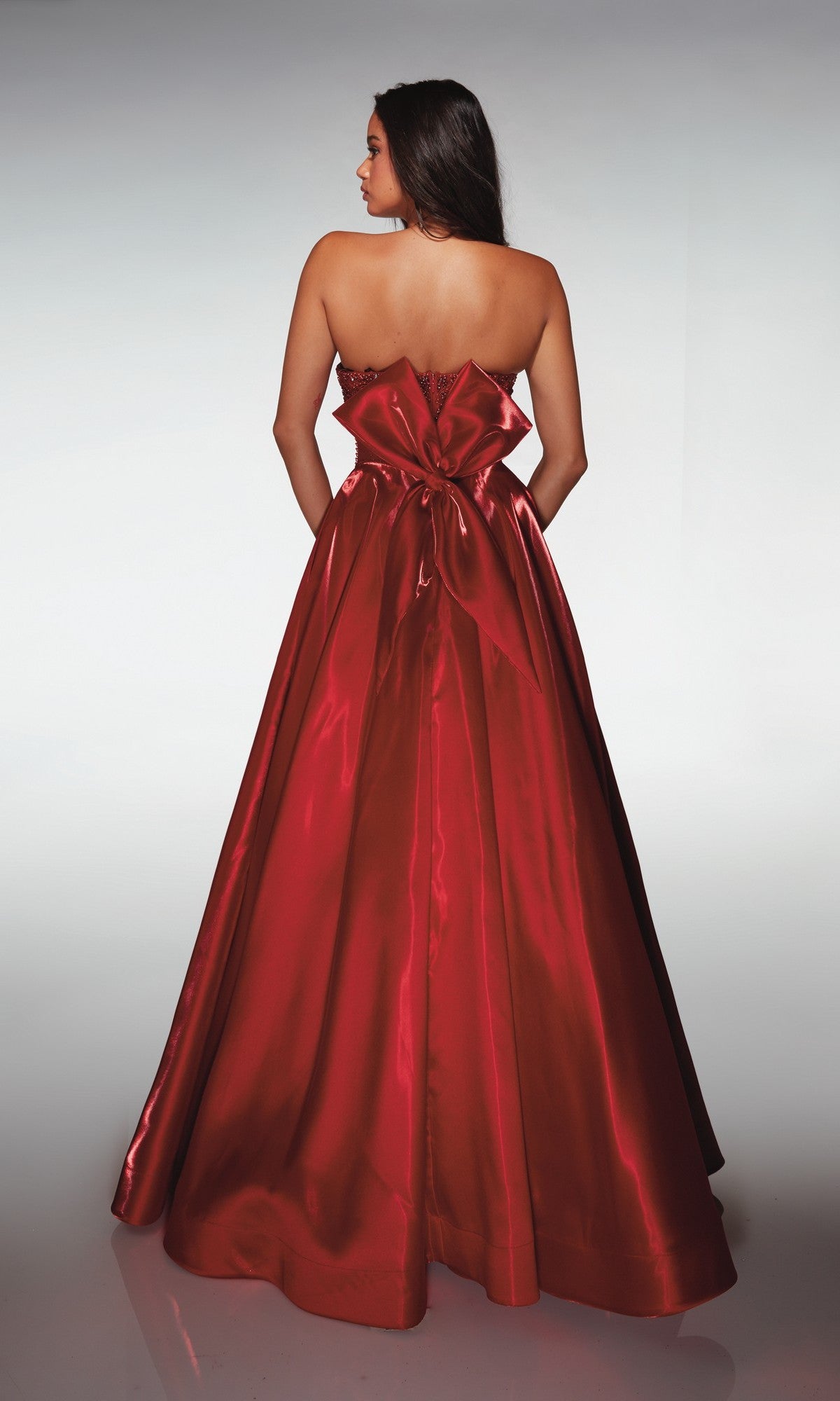A woman stands with her back to the camera, wearing the floor-length Alyce 62134 Long Prom Dress—a strapless satin ball gown in shiny red with a large waist bow.